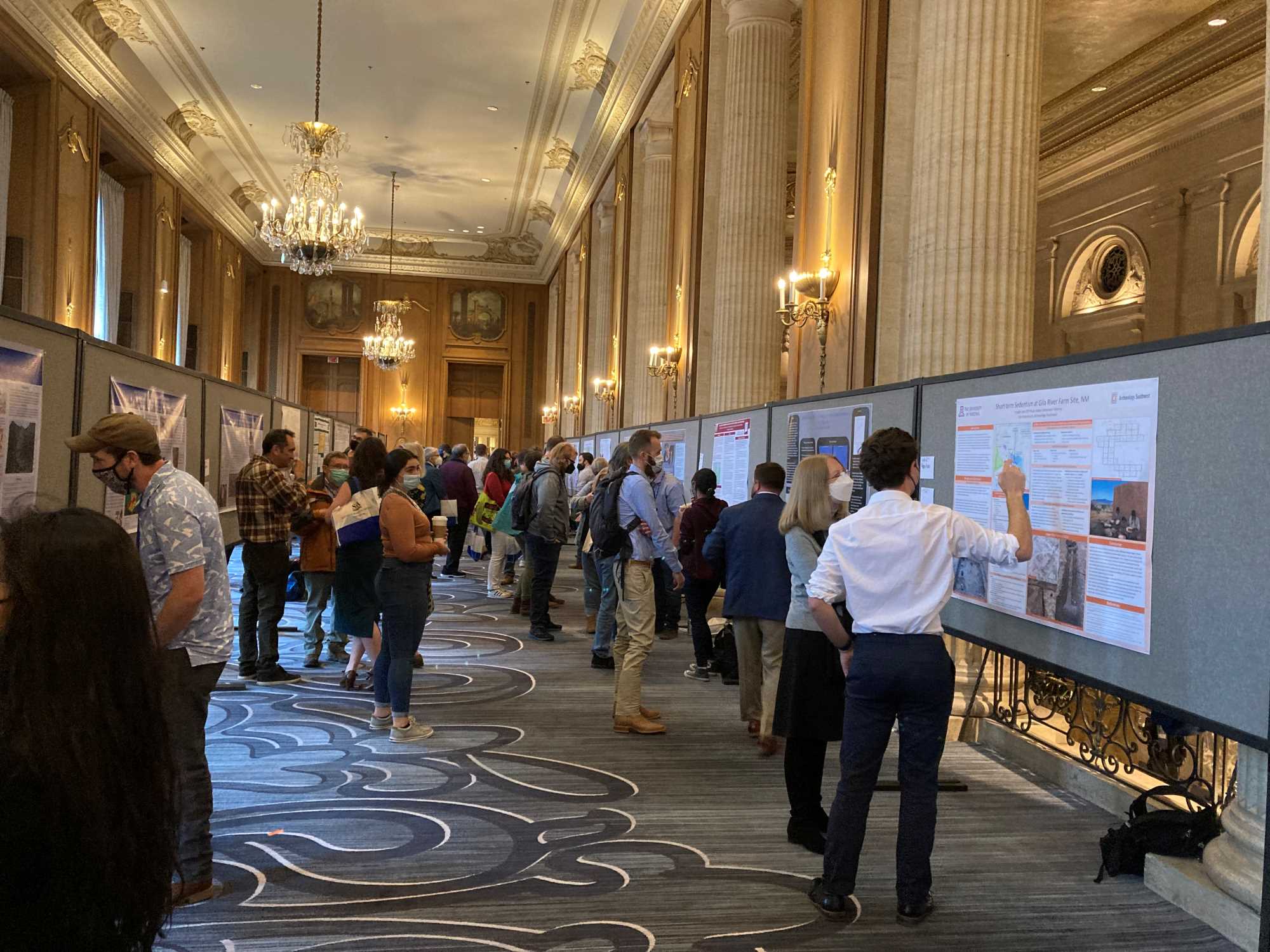 This image is taken from one end of a long, high-ceilinged gallery. Along wither side of the gallery are temporary bulletin boards where people are presenting posters to groups of meeting attendees.