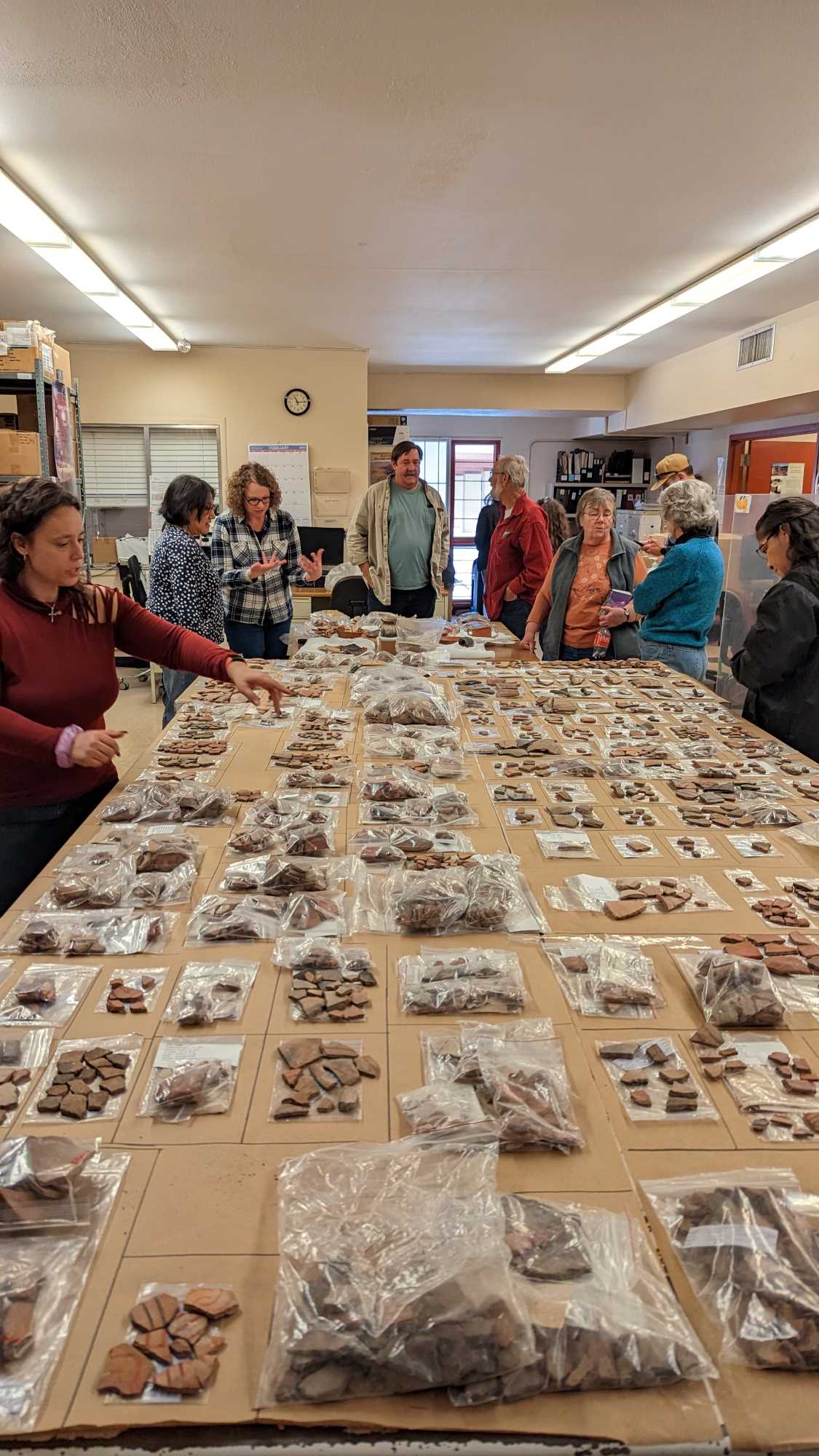 A large group of people is in a lab gathered around tables. The tables display groups of pottery sherds and other artifacts.