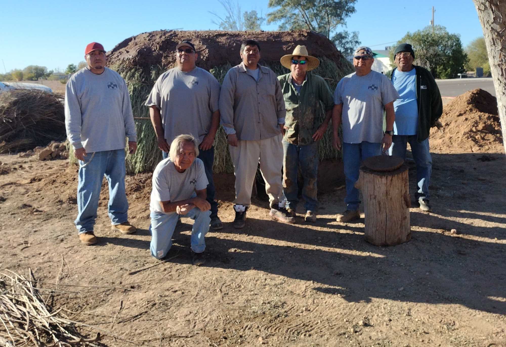 Six men stand shoulder to shoulder in front of the remodeled pithouse. The new roof mud is on and fresh arrowweed thatching surrounds the exterior. Another man kneels just in front of the group.