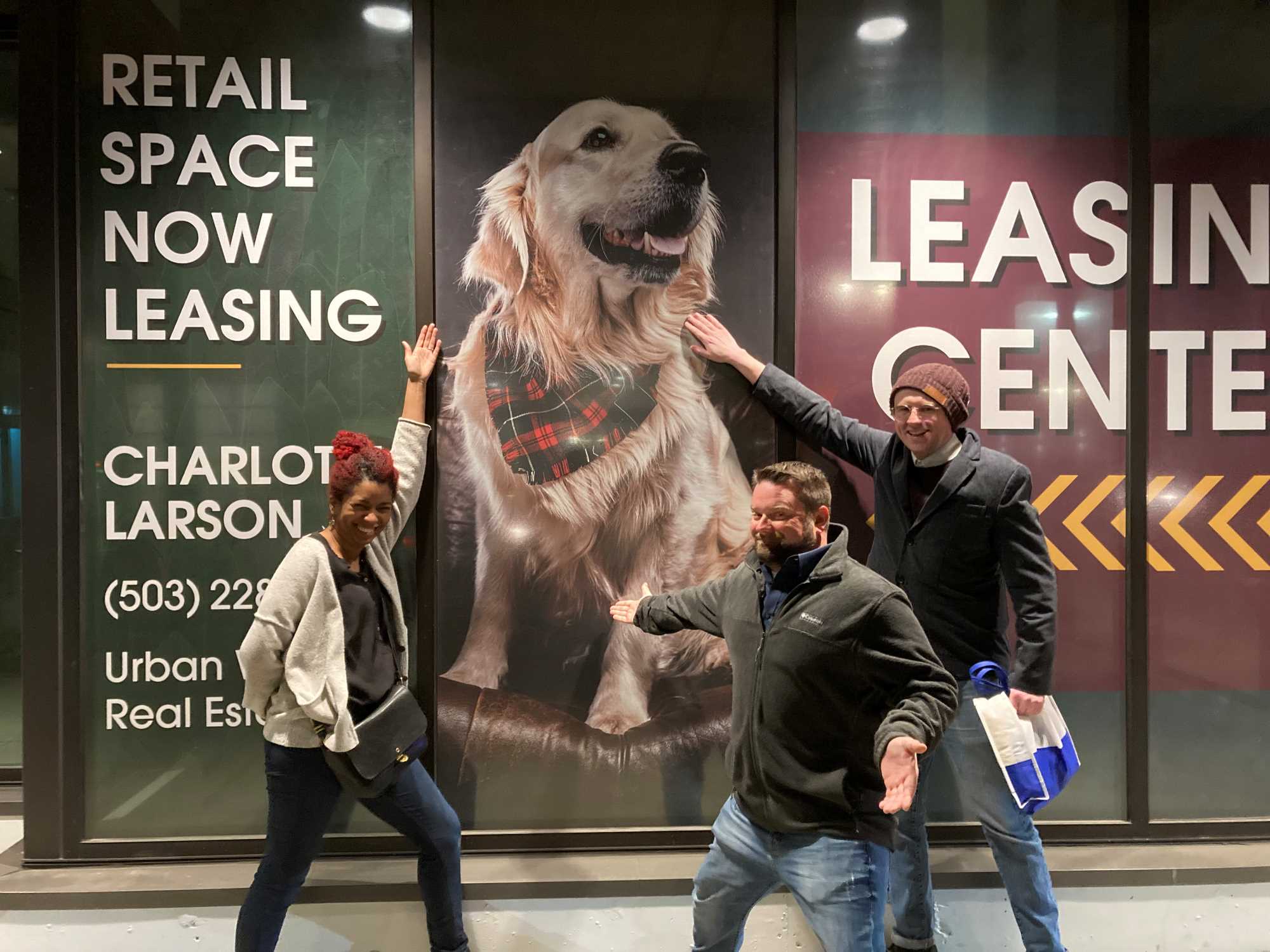 Three people are goofily gesturing to a large poster of a golden retriever.