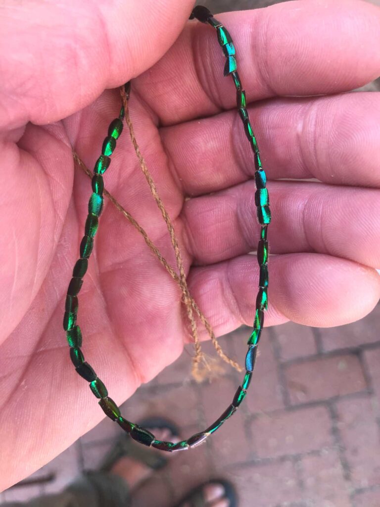 A string of iridescent green beads made from the hind legs of fig-eater beetles and strung onto natural fiber cordage is held in the palm of a human hand.