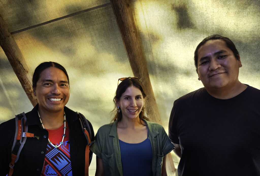 Three people are standing inside a large canvas tent. From left to right they are Indigenous conservationists and scholars Angelo Baca (Dine), Ashleigh Thompson (Red Lake Ojibwe), and Skylar Begay (Dine, Mandan and Hidatsa)