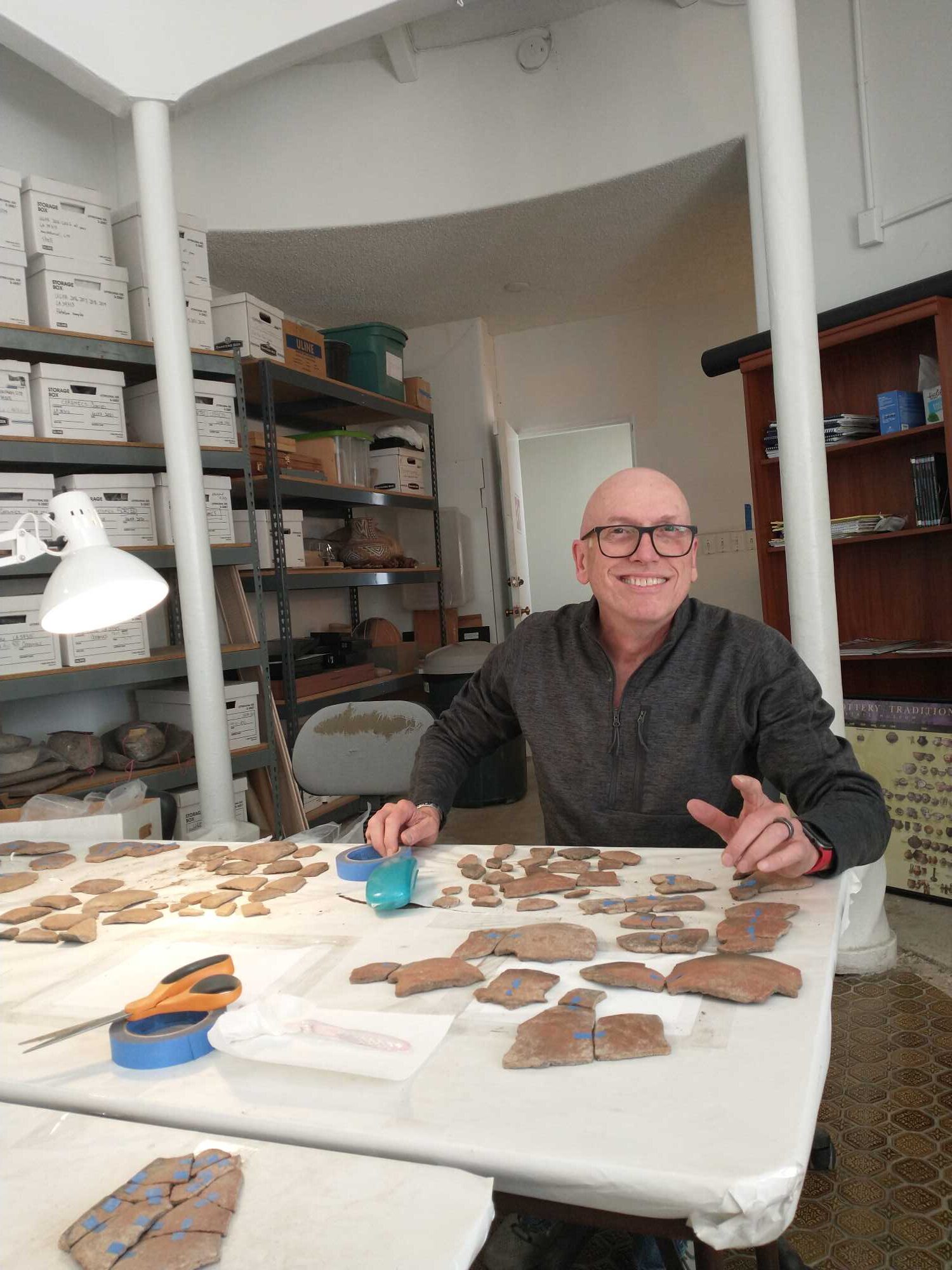 A ma is sitting at a table in an archaeological laboratory. He is using blue masking tape to join pottery sherds that fit together.