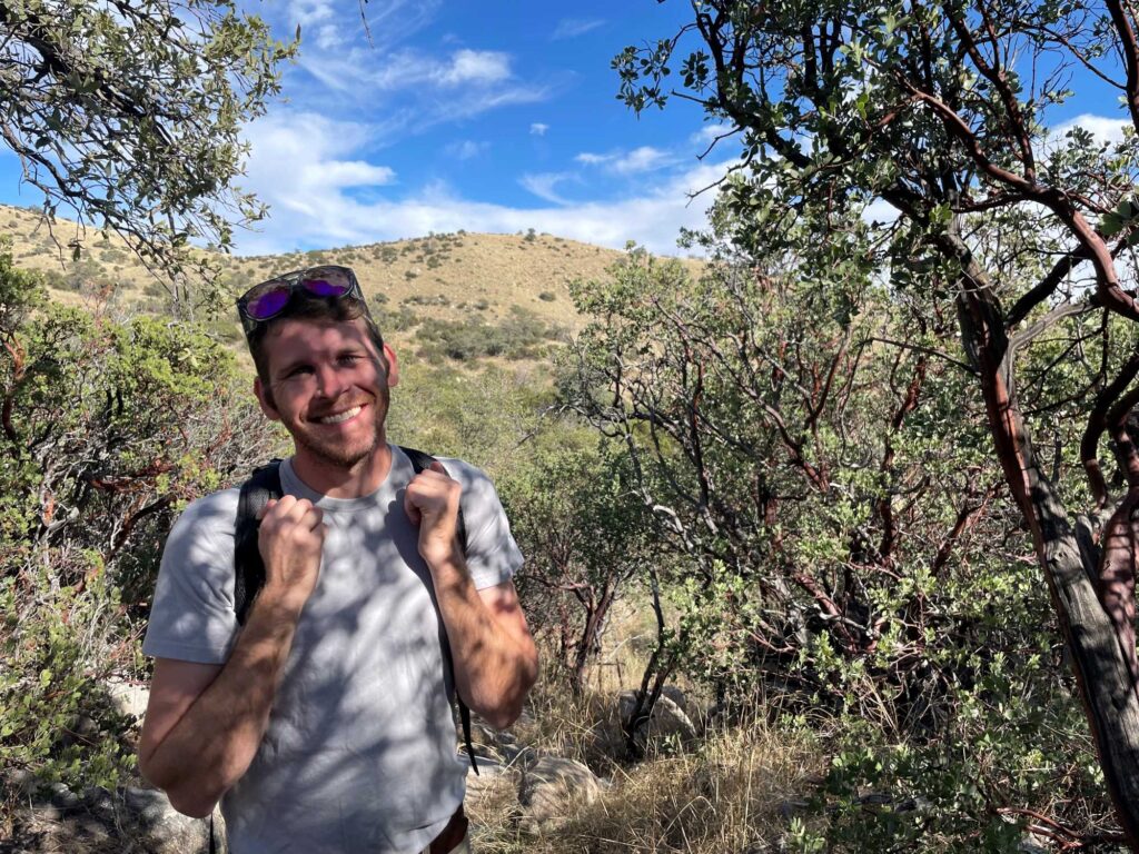 A young adult man is standing outdoors and shaded by trees. He has a backpack and his sunglasses are on top of his head. There is a mountain foothill in the background.