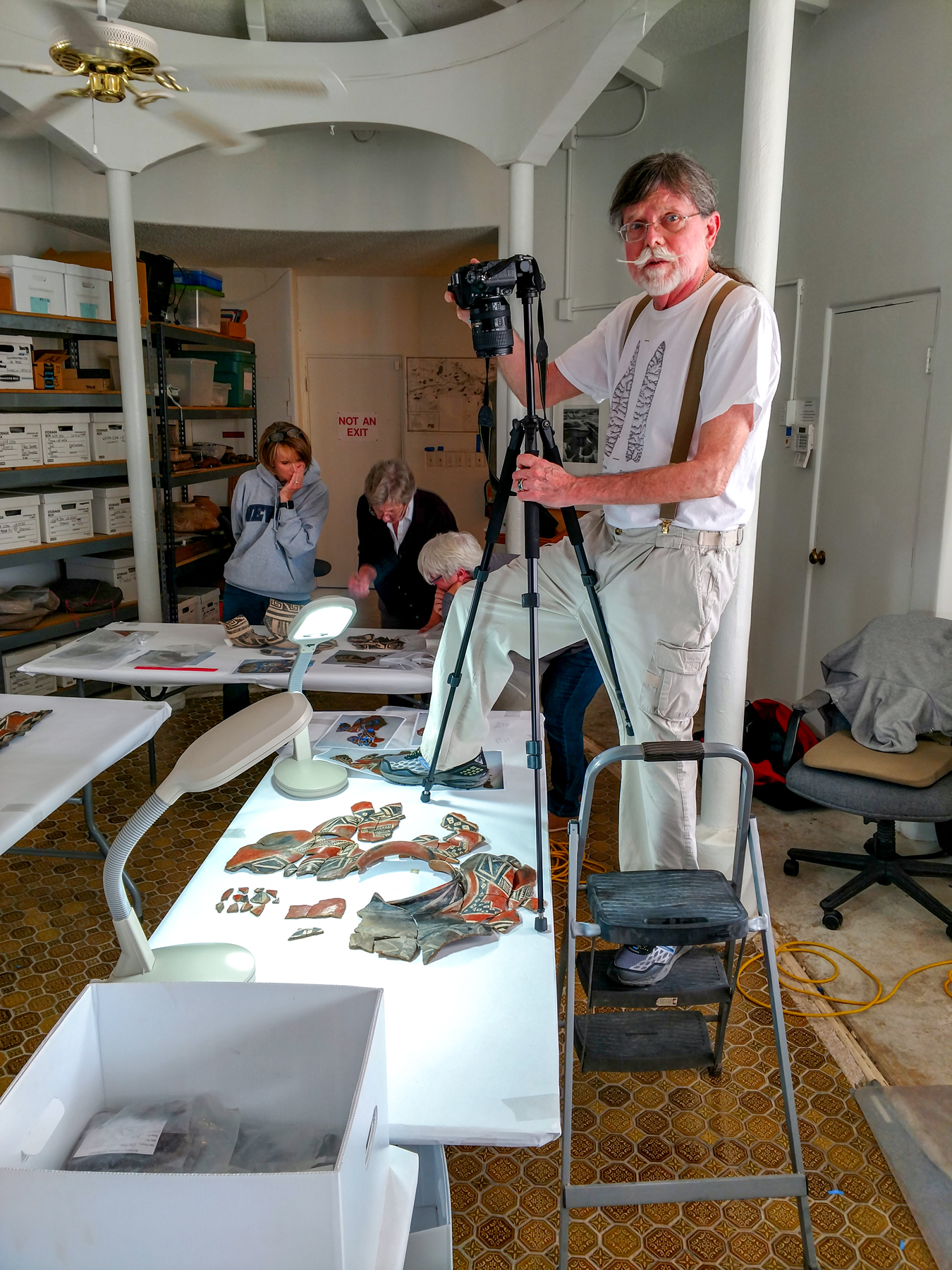 A man stands on a step stool above a table in an archaeological laboratory. He has a camera rig and is taking photos of artifacts from above. Three people are working in the background.