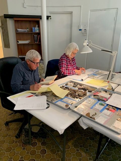 A man and a woman are working at a table in an archaeological laboratory. They have trays of artifacts in front of them and they are taking notes on legal pads. A lamp illuminates their work from above.