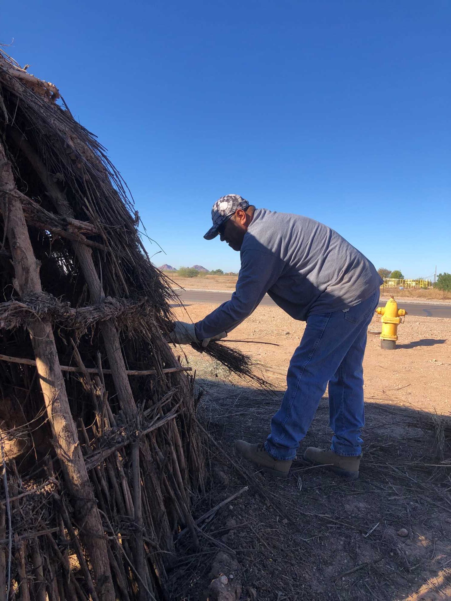 A man stands outside the pithouse to the right. He is bent at the waist and removing old arrowweed from the support structure.