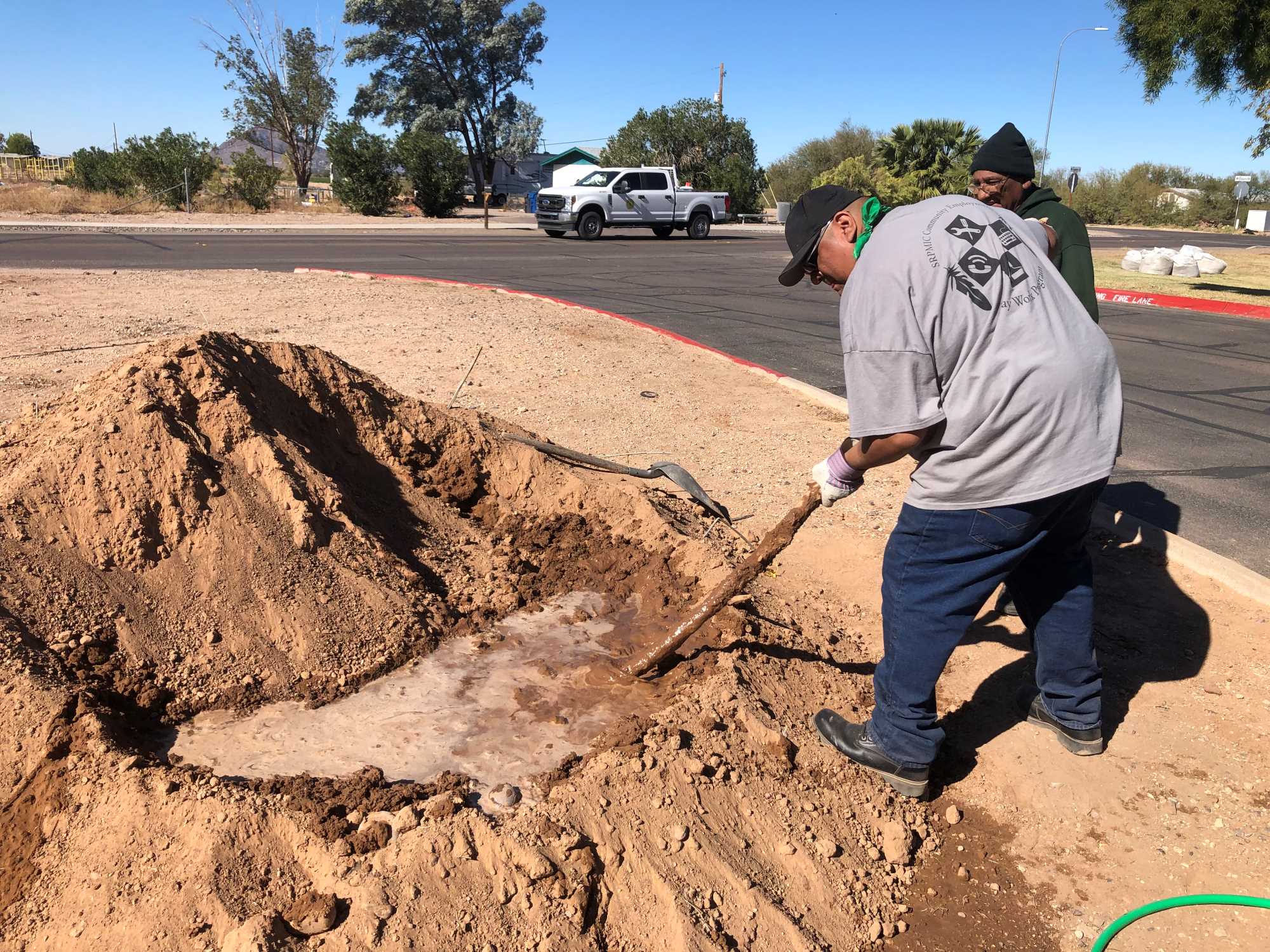 To the right of the mud pit, a man is slightly bent and using a wooden pole to mix the clay with the water. Another man is standing to his right.