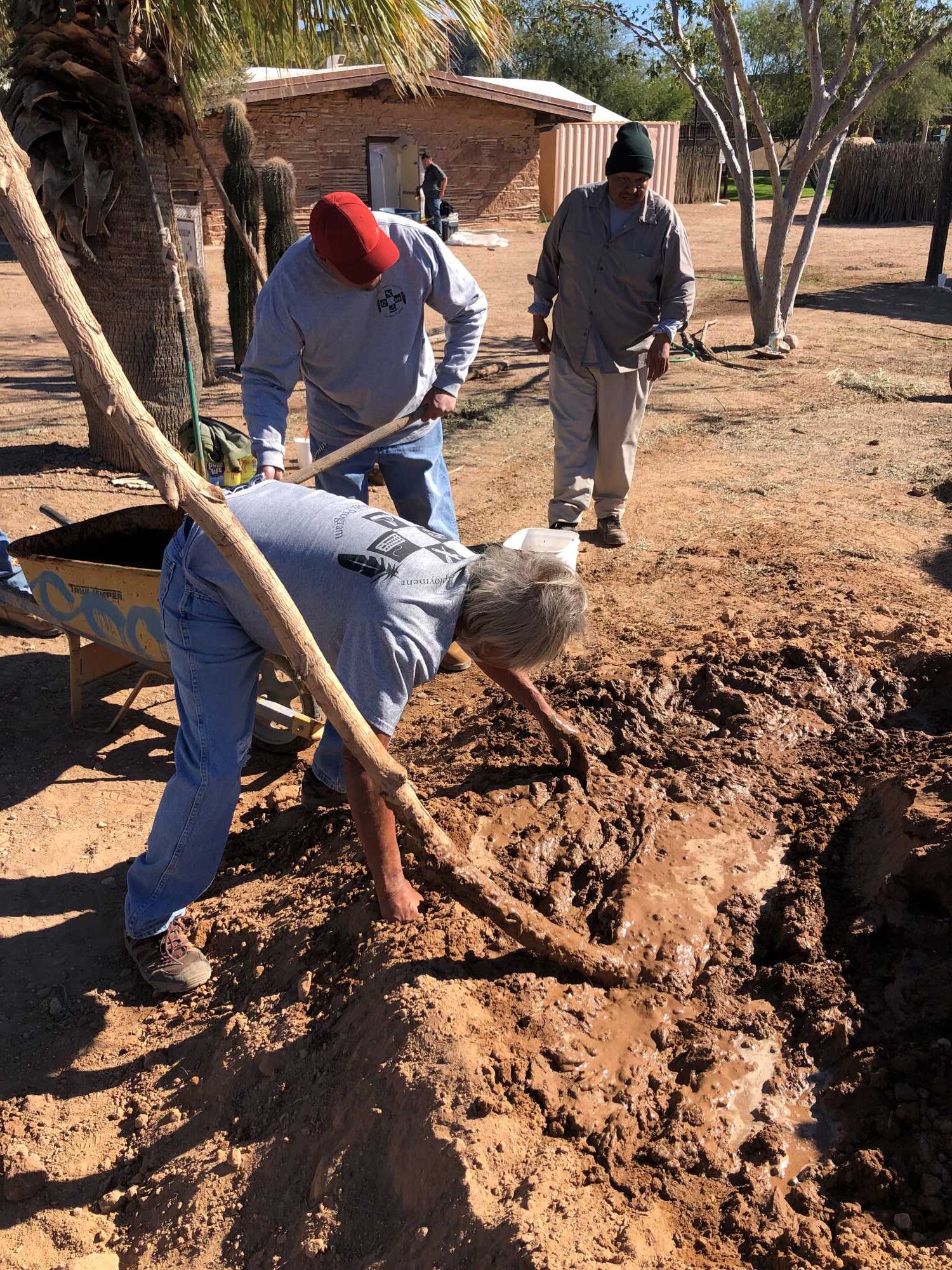 The person in the foreground is bent at the waist over a large muddle pit. He is mixing the mud with his hands. The person in the middle ground is shoveling clay from a wheelbarrow into the pit. A third person is approaching from the back.