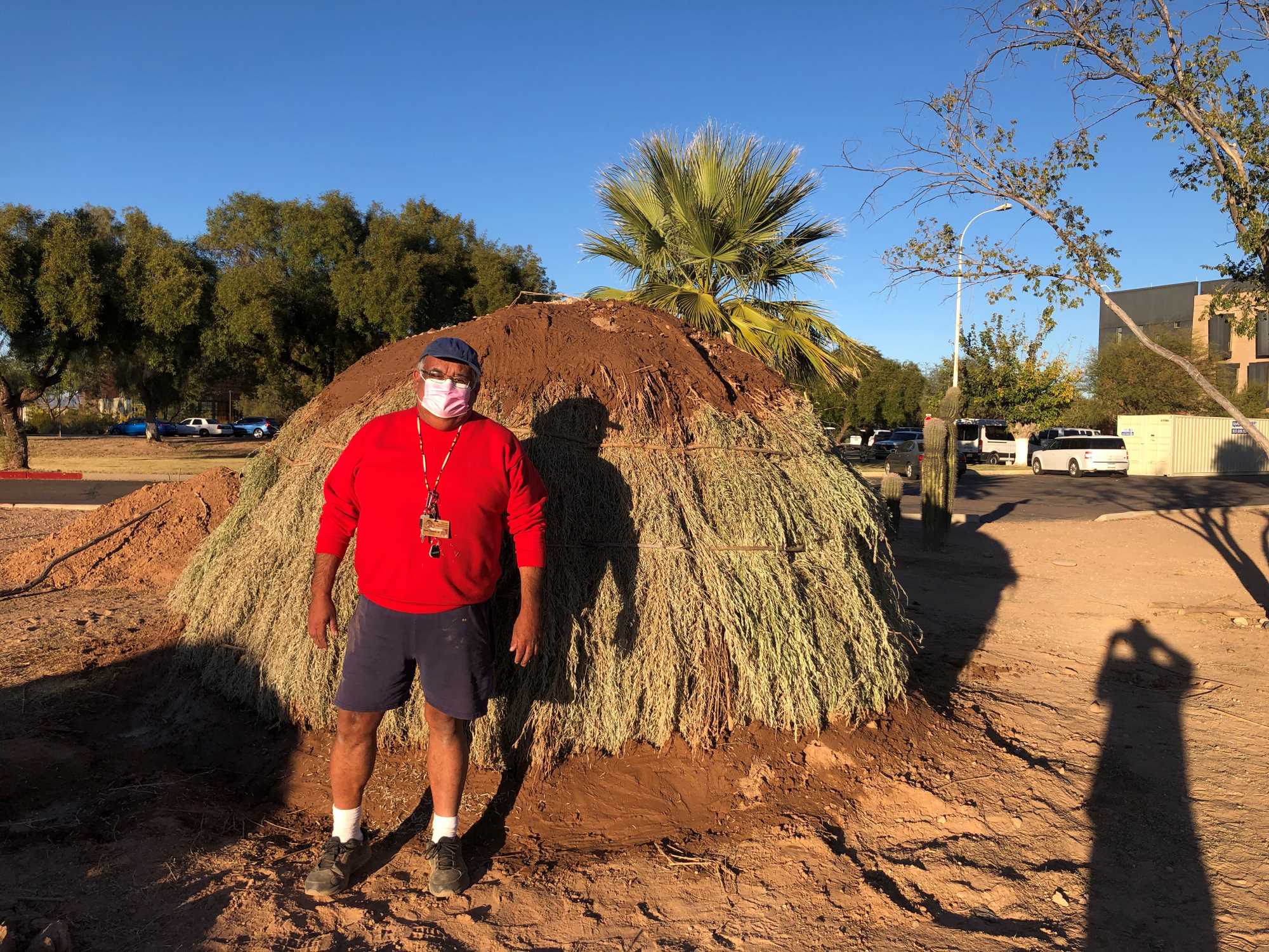 The museum director stands in front of the fully remodeled replica O'odham pithouse. He is wearing a red sweater, ballcap, and face mask.