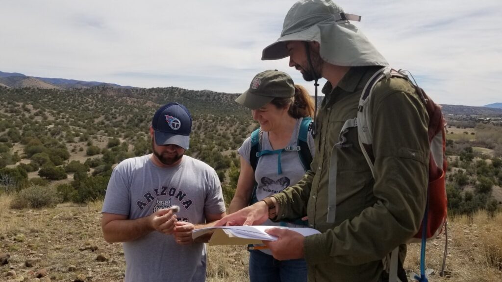 Three people are in the frame against a backdrop of desert mountain foothills with scrub, grasslands, and juniper. At left is a man in a baseball cap holding a tuft of fur for examination by his solleagues, a woman in a ball cap in the middle and another man at fight in a sun hat and holding paperwork.