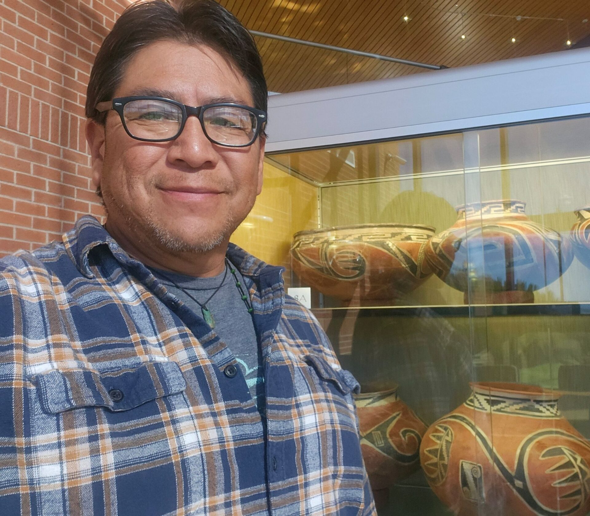 A portrait of Hopi archaeologist and river guide Lyle Balnquah. Lyle is smiling into the camera for a selfie and is standing next to a museum display of ancestral pottery with exterior colors of red, black, and white.