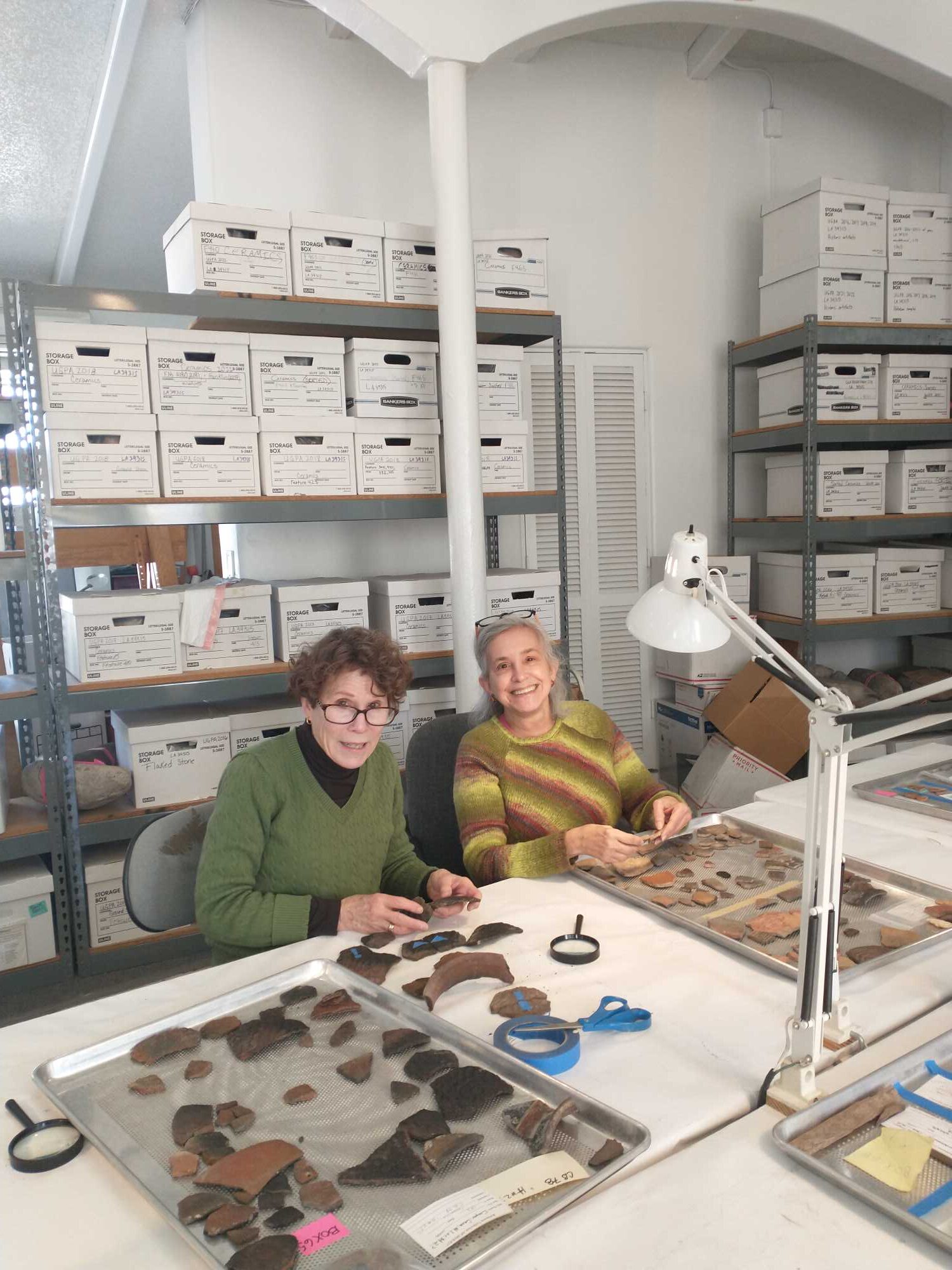 The image shows two women volunteers examining and sorting pottery sherds at a table in an archaeological lab. Behind them is shelving full of museum collection storage boxes. 