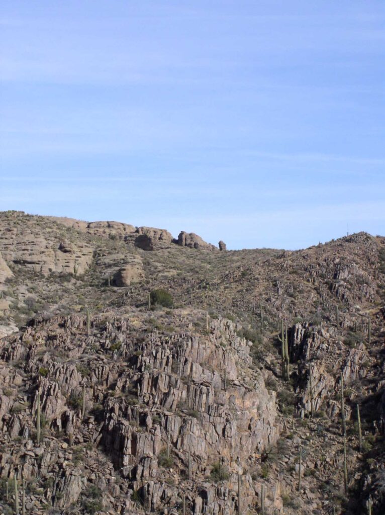 This image shows an ancient foot trail in the mountains of southern Arizona. The landscape is rocky and there are saguaro cacti. A blus sky rises above.