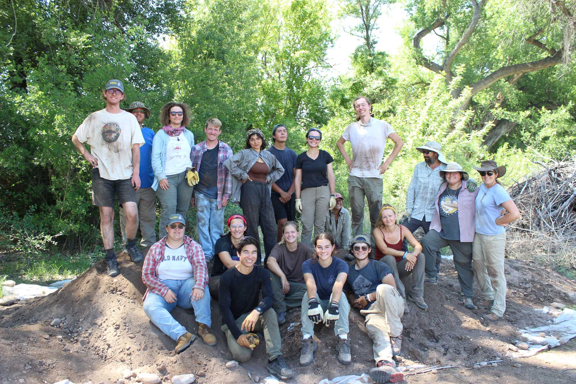 This image shows a group of archaeology field school students, dirty and happy after backfilling the trenches to prevent erosion and damage. There are two rows, one standing in the background, and one seated in the foreground.