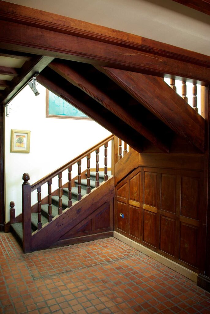 This image shows a solid wooden staircase with wooden railings and wooden paneling on the interior of a mansion. The floor comprises small terracotta tiles.