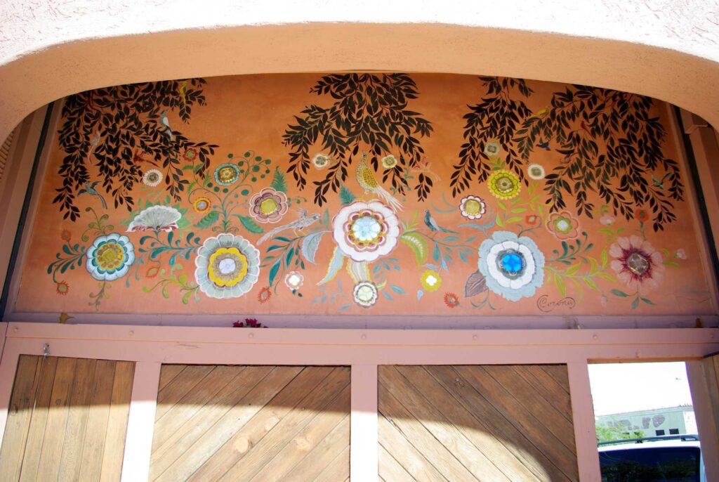 This image shows a mural above a large wooden exterior gate at the entrance of the Bates Mansion. The motifs include dark green vines and colorful flowers.