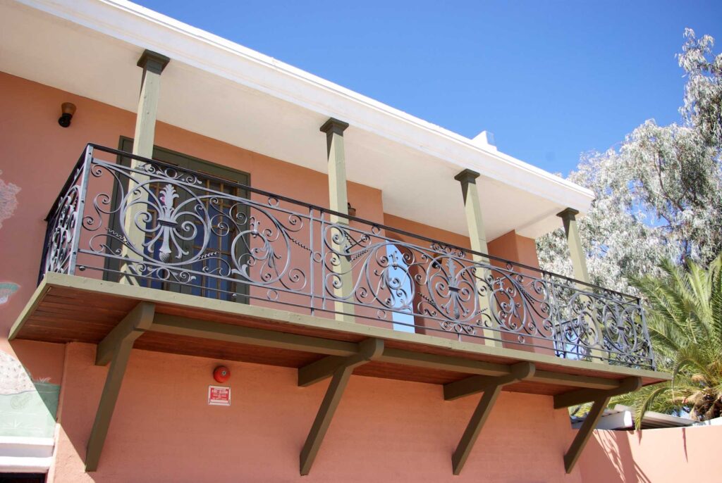 This image shows an exterior second-story wrought iron balcony at the Bates Mansion. The exterior adobe wall is a pinkish-terracotta color. The wrought iron motifs include scrolls and medallions.