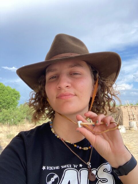 Image shows a woman wearing the wool felt fedora she has added a chin strap and a pendant to. The pendant helps adjust the tightness of the strap and is made of ground stone.