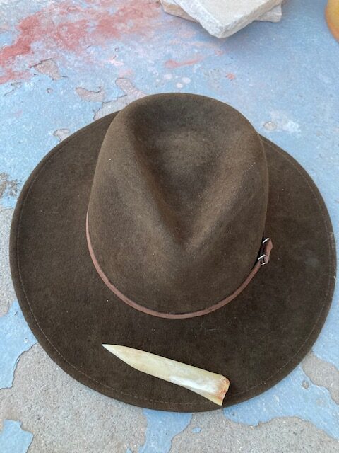 Image shows a wool felt fedora from above with a bone awl resting on the brim. The awl was used to create holes for a chin strap.