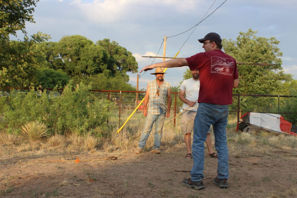 A man in a red t-shirt and black ball cap stands at the right side of the frame. His back is to the viewer. His left arm is extended to sight the atlatl and dart he is ready to project with his right hand.