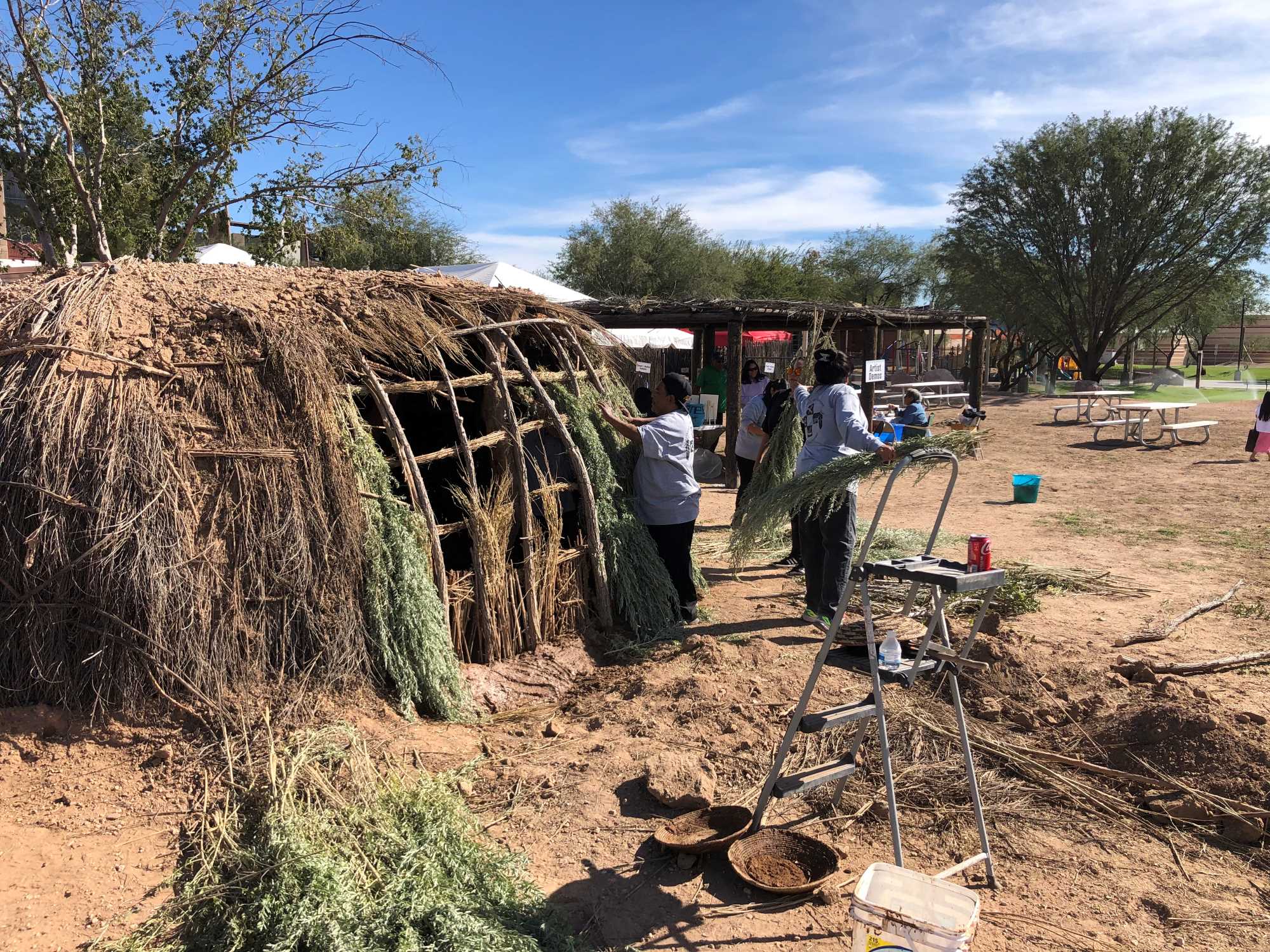 One person is weaving fresh arrowweed into the exterior of the pithouse. A second person stands nearby holding two more bundles of arrowweed.